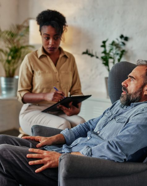 An attentive african female counselor listens and takes notes as a vulnerable middle age male patient discusses an important topic.